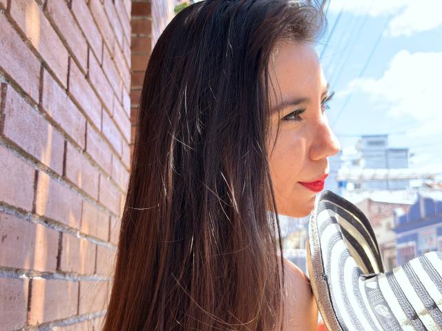 FreyaraVenwo with long brown hair, red lipstick, and striped hat, leaning against a brick wall, sunlight on her face, looking to the side, blue sky in the background.