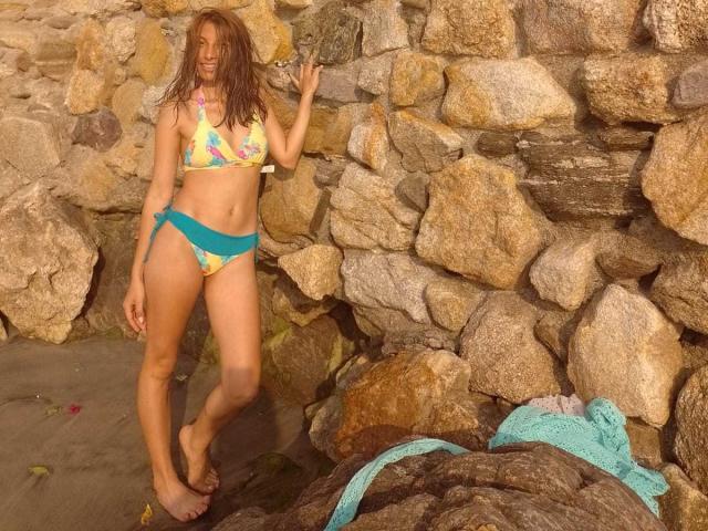 KiaraWiild se tient pieds nus sur une plage rocheuse, vêtue d'un bikini jaune à motifs floraux avec des bretelles bleues, tenant un rocher, avec les cheveux bruns mouillés, et une serviette turquoise sur les rochers..