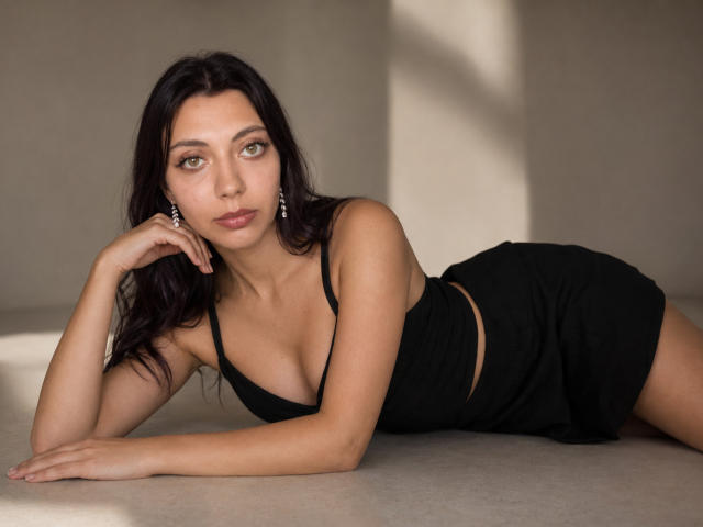 SammieZera, avec ses cheveux foncés, ses yeux verts et son joli visage, allongée sur le côté en robe noire, montrant ses seins et ses fesses, portant des boucles d'oreilles pendantes, dans une lumière douce..