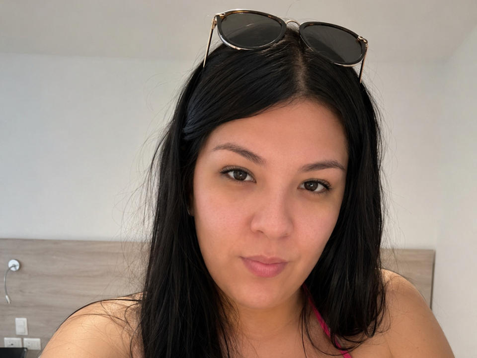 NanceLuci hat schwarzes Haar, braune Augen und einen hellen Hautton, trägt eine Sonnenbrille auf dem Kopf und rosa Träger über der Schulter und zieht leicht die Lippen zusammen..
