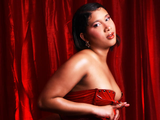 LoreDuval, avec des lèvres rouges brillantes, portant un corset rouge en cuir sans bretelles, ses seins galbés partiellement visibles, tenant les côtés, cheveux noirs courts, fond de rideau rouge, expression sensuelle..