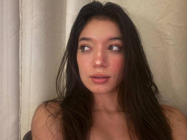MichellFord has long, dark hair, pinkish-red eyes, and slightly parted lips. She's looking to the side with a subtle, pouty expression. Her nose is small, and she's topless.