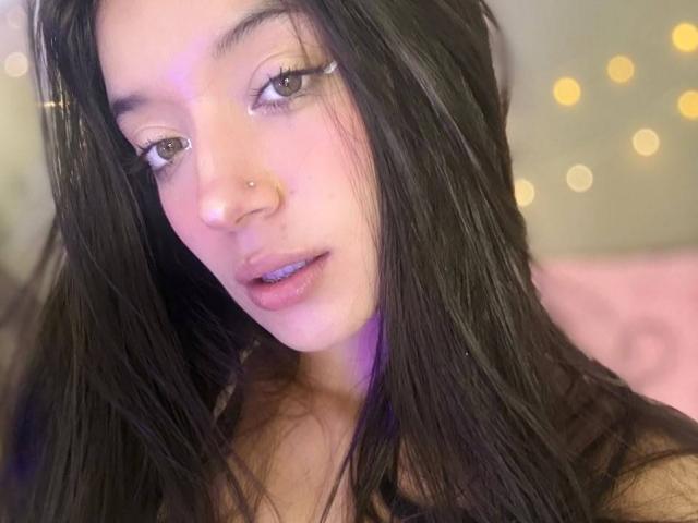 MichellFord tem cabelos pretos longos, um piercing no nariz e batom azul. Ela está olhando para a câmera com os olhos semicerrados, usando delineador preto. Seu rosto está levemente avermelhado..