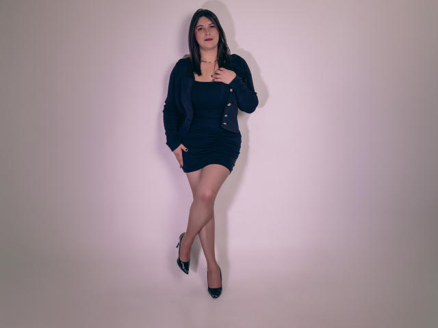 ZoeyGrey, com cabelo escuro, vestindo um cardigã preto e um vestido curto, em pé contra uma parede lisa, com as pernas cruzadas, saltos altos pretos, a mão no peito, olhando para cima..
