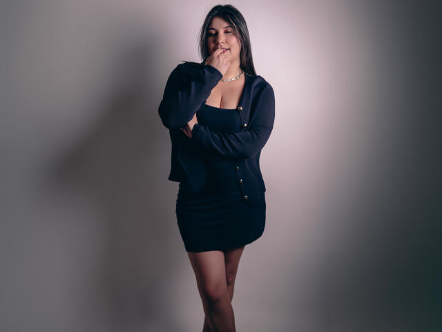 ZoeyGrey está de pie con una sonrisa tímida, la mano cerca de su boca, vestida con un vestido y una chaqueta azul marino, que resaltan sus pechos debido a su ajuste ceñido, frente a una pared gris..