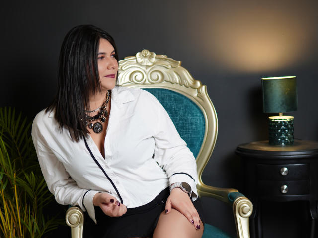 ZoeyGrey, avec des cheveux noirs, une chemise blanche à boutons, une jupe noire, un collier et une montre, assise sur une chaise ornée, regardant sur le côté, avec du vernis à ongles noir, une lampe verte sur une table sombre..