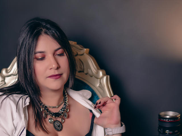 ZoeyGrey with dark hair, wearing a white jacket, holding a cigarette, looking down, pink eye makeup, necklace with large beads, black bra, camera on right.