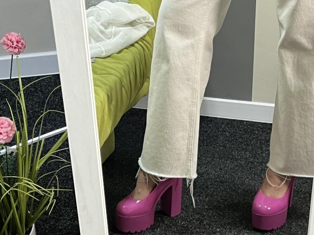 Valnes' roze plateauhakken met dikke zolen, witte, versleten denim, en blote voeten die op een donker tapijt staan, worden weergegeven in een spiegel met roze bloemen aan de linkerkant..