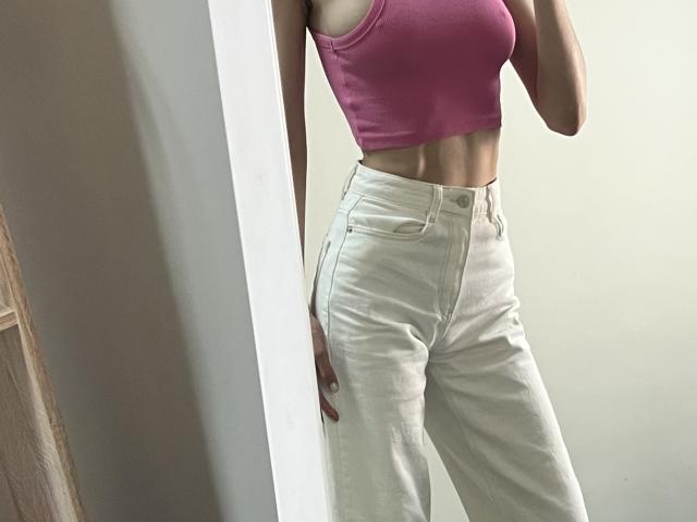 Valnes indossa una canotta rosa che mette in mostra il seno, e pantaloni bianchi a vita alta, in piedi accanto a uno specchio, mostrando il suo ventre piatto e i muscoli addominali tonici..