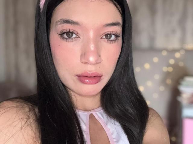 MichellFord has long black hair, pinkish skin, brown eyes, and glossy pink lips. She wears a pink headband, and a white halter top with a keyhole cutout.