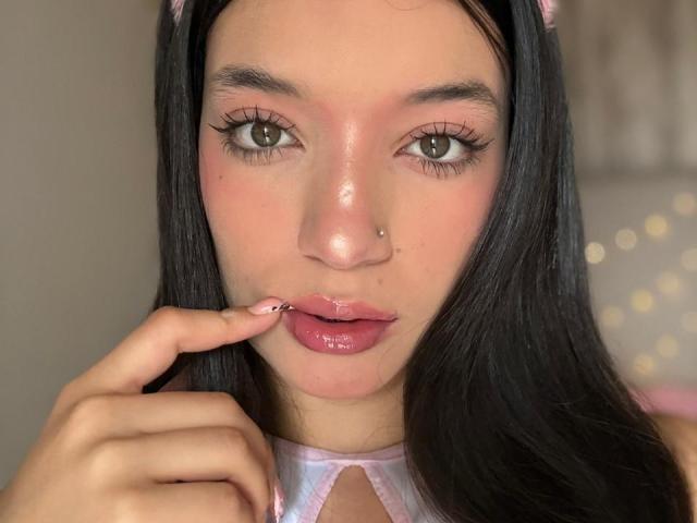MichellFord con labbra rosa lucide, occhi marroni, capelli neri, fascia rosa per capelli, piercing al naso, che tocca le labbra con un dito, indossa una maglietta rosa e bianca, e guarda la fotocamera..