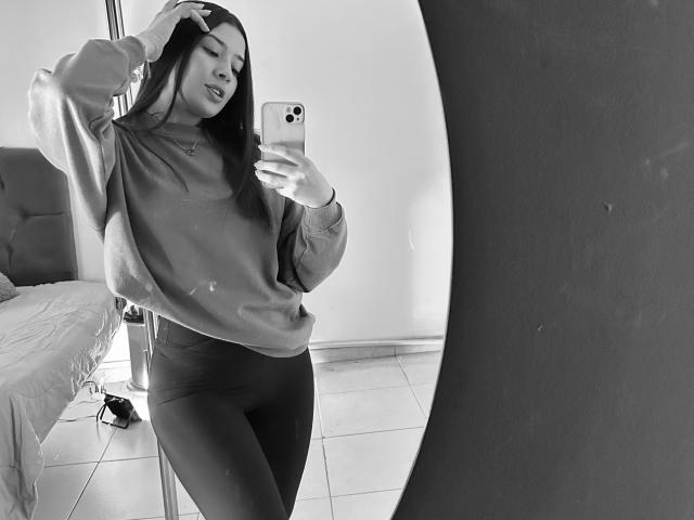 EmmaLombardi with long dark hair, wearing a loose sweater and tight pants, taking a mirror selfie, hand in hair, showing her ass in a black-and-white photo.