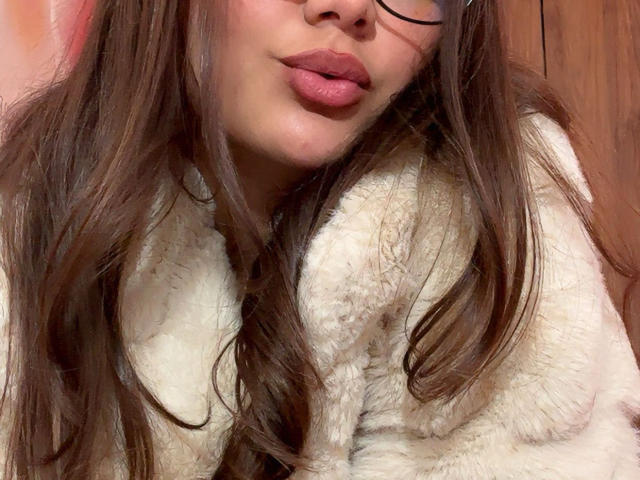 DianaFernandezz con labbra rosa e piene, lunghi capelli castani, che indossa occhiali e un cappotto bianco soffice. Il seno è parzialmente visibile, la texture del cappotto è morbida. Sfondo in legno. Immagine fotografica..