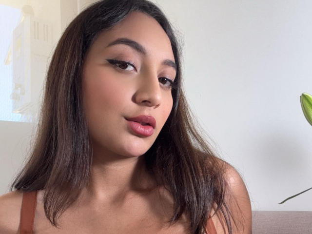 DianaFernandezz ha lunghi capelli castani, occhi castani e rossetto rosa. Indossa una cinghia marrone, guarda leggermente di lato con un sorriso sottile e seducente..