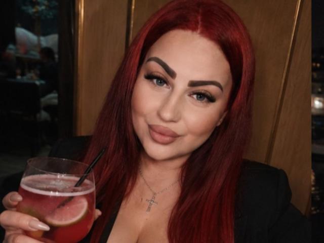 MyraKeldy met rood haar, die een drankje vasthoudt met een rietje en een citroenschijfje, gekleed in een zwarte top en met een kruisbeeldketting, lachend naar de camera..