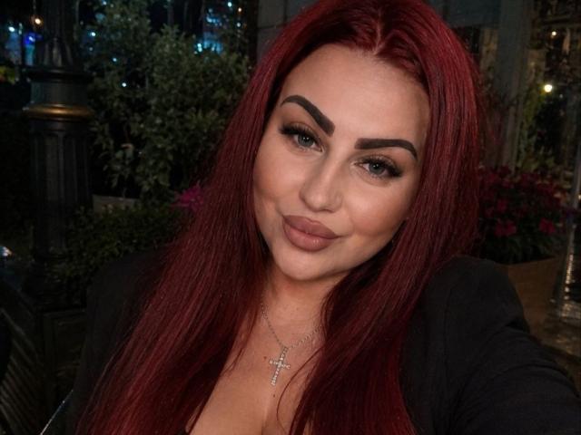 MyraKeldy tem cabelo ruivo, sobrancelhas grossas e lábios cheios com um leve sorriso. Ela está vestindo uma blusa preta e um colar com uma cruz, com um sorriso sexy e confiante..