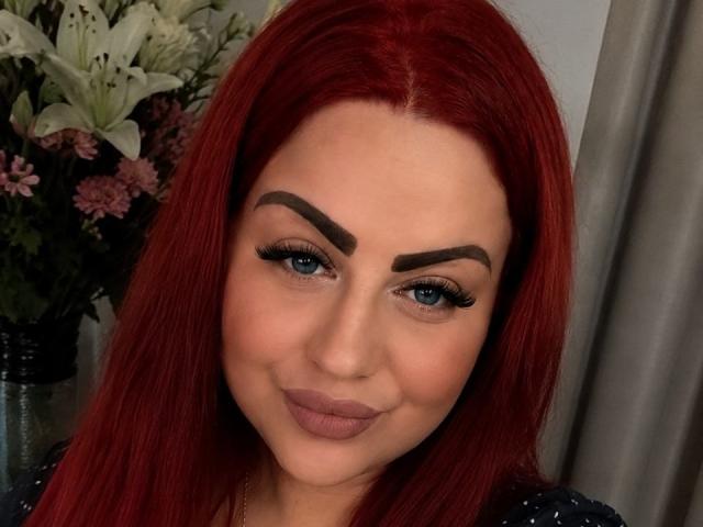 MyraKeldy heeft rood haar, blauwe ogen en dikke wenkbrauwen met lange wimpers. Ze glimlacht met roze lippenstift, waarbij haar volle lippen zichtbaar zijn. Haar haar is recht en ze kijkt naar de camera..