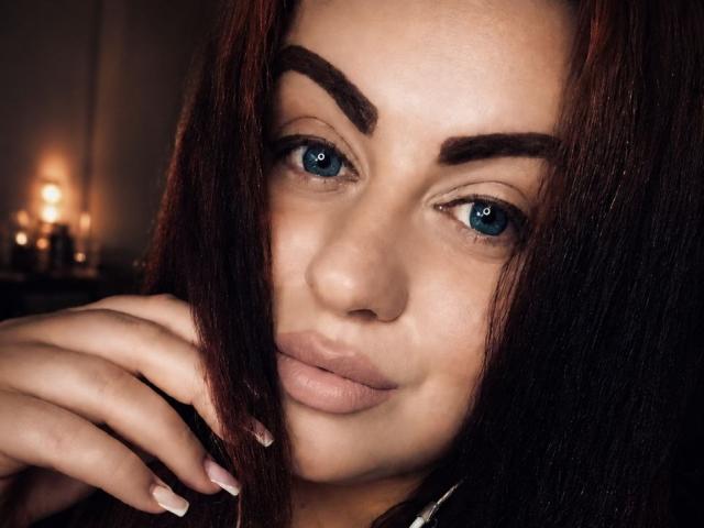 Le visage de Myra Keldy, avec ses sourcils marron foncé, ses yeux bleus et sa peau brun clair. Elle a les cheveux brun rougeâtre, légèrement séparés, et sa main touche sa joue. Ses ongles sont manucurés..