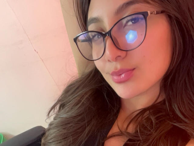 DianaFernandezz con gli occhiali, capelli castani ondulati, labbra rosa lucide e un riflesso blu nella sua pupilla destra, indossa una maglietta nera e guarda verso la fotocamera..