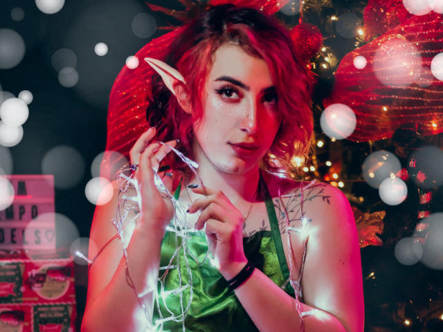 Un gato con cabello rojo, orejas de elfo y pecas, sosteniendo luces de Navidad, vistiendo una blusa verde, en una escena festiva y brillante. Se ven visibles los senos, el trasero y los tatuajes..