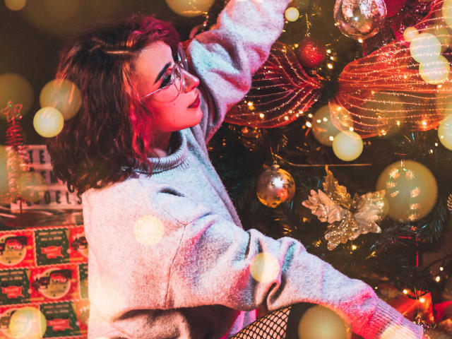Een kat met een bril, krullend bruin haar en een grijze trui, die reikt naar kerstversieringen. Bokeh-lichten, rode en gouden decoraties en een glinsterende boom op de achtergrond..
