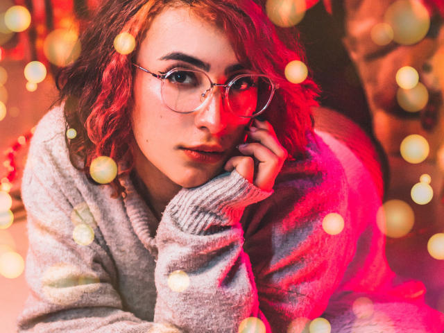 A CatsitoS tem cabelo ruivo encaracolado, usa óculos e um suéter cinza, está deitada com uma luz vermelha e efeito bokeh, com a cabeça apoiada na mão..