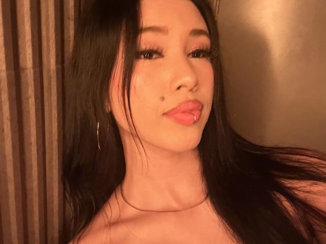 NashiroSakuraii has long black hair, a small mole on her left cheek, and is pouting with pink lips, wearing a thin necklace and small hoop earrings, in a warm light.