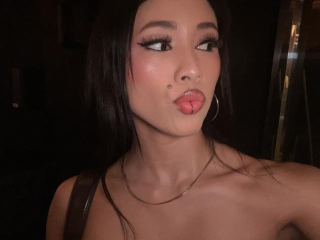 NashiroSakuraii with dark hair, pouty pink tongue out, heavy eyeliner, small mole on nose, gold hoop earrings, light skin, no clothes, subtle makeup, slight sheen on face.