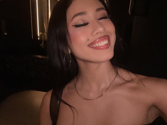 NashiroSakuraii with dark hair, closed eyes, and red lipstick, smiling with tongue out, showing small mole on left cheek, wearing hoop earrings, and thin necklace, in dim lighting.