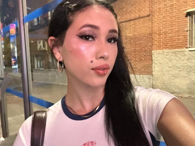 NashiroSakuraii with glossy pink lips, dark braided hair, and a small mole on her left cheek, wearing a white shirt with blue trim, standing by a glass barrier.