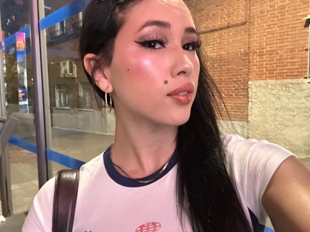 NashiroSakuraii has shiny pinkish skin, thick black eyebrows, long eyelashes, a small beauty mark on her left cheek, and glossy pink lips. She's wearing a white tee with blue trim.