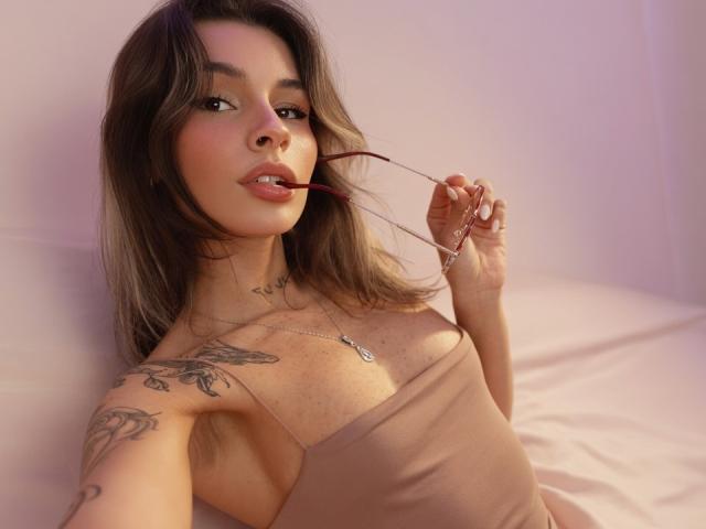 LenaBranch with brown hair, wearing a beige tank top, holding red glasses in her mouth, showing tattoos on her arm and chest, looking at the camera.