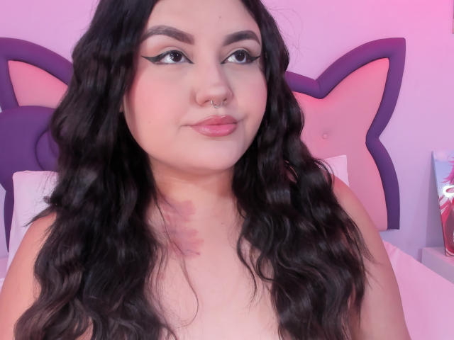 MarlaBriard with long, wavy black hair, nose ring, and eyeliner, smiling with pink lips, topless, showing a red mark on her chest, in a pink-lit room.