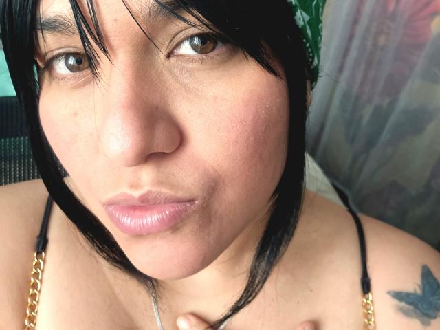 VivianaThompson has brown eyes, black hair with bangs, and pinkish lips. She's wearing a gold chain necklace, and her left shoulder has a blue tattoo.