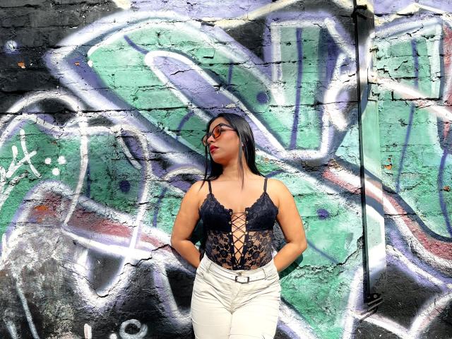 MelodyRoss with long black hair, wearing glasses, a black lace-up bustier, and white pants, standing against a graffiti-covered wall, hands in pockets, looking to the side.