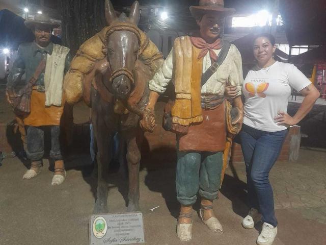 HypnoticBella stands next to a life-sized bronze statue of a cowboy with a horse. She's wearing a white shirt with an orange fruit logo and blue jeans.