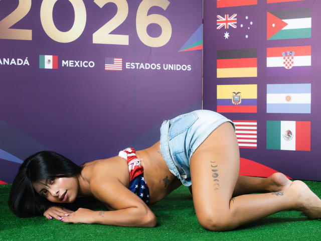 LeiaLaurent está deitada na grama, em posição de quatro, vestindo shorts de jeans e uma blusa sem alças nas cores vermelho, branco e azul. Ela tem tatuagens na coxa esquerda e cabelo preto..