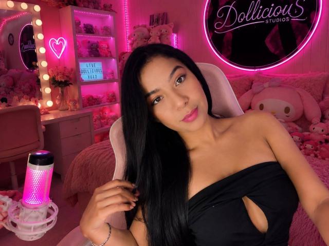 Yasmine Tapias, con lunghi capelli neri, rossetto rosa e un abito nero con tagli, seduta in una stanza illuminata di rosa, che si tiene i capelli, con un cuore neon rosa e una lampada viola..