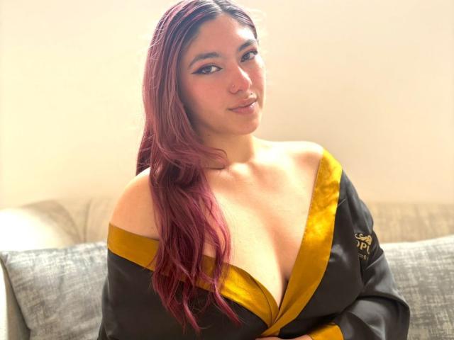 DaphneLenoir, con i capelli rossi, indossa una veste nera e dorata che lascia intravedere il seno, seduta su un divano grigio, sorride dolcemente alla telecamera..