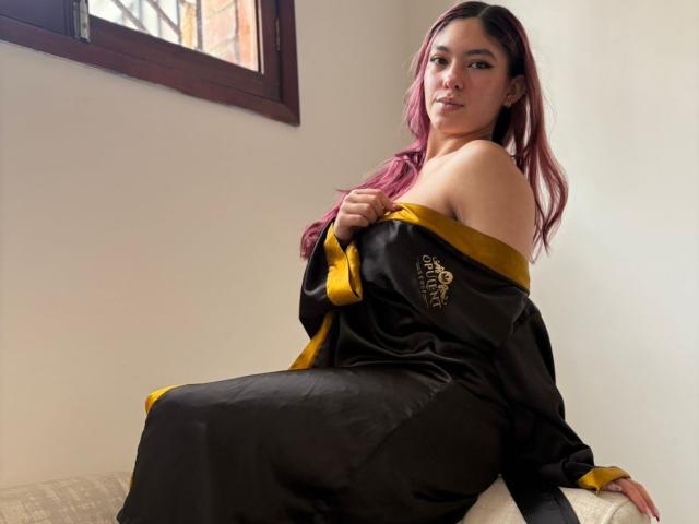 DaphneLenoir com cabelo rosa, robe de cetim preto e amarelo, com um ombro descoberto, sentada no sofá, olhando por cima do ombro, segurando o robe, revelando o ombro nu, fofa, sexy, luz suave..