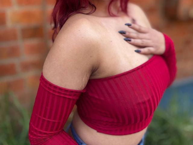 DaphneLenoir portant un haut rouge à rayures, avec une épaule dénudée, une main sur la poitrine, montrant ses seins, des cheveux roux et un vernis à ongles foncé, debout devant un mur de briques..