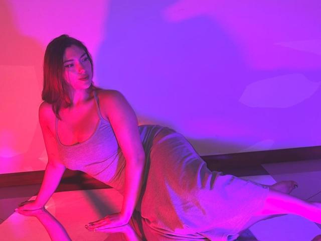 DaphneLenoir, ronde avec de grandes seins, est allongée dans une lumière rose et violette, vêtue d'un débardeur gris scintillant et d'une jupe assortie, appuyée contre une surface blanche, et elle sourit..