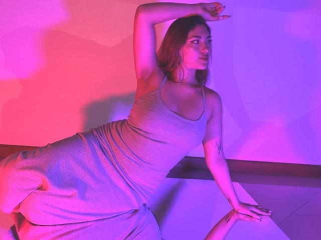 DaphneLenoir liegt mit einem Arm erhoben da und trägt ein eng anliegendes, graues Kleid, das ihr Gesäß und ihre kleinen Brüste zur Schau stellt, wobei rosafarbenes und violettes Licht verwendet wird..