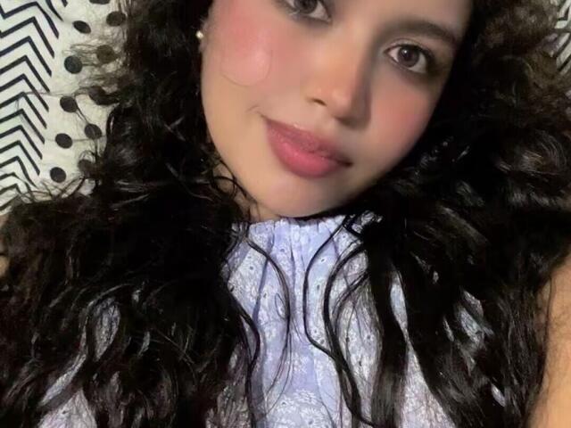 JeimyCampos with curly black hair, light skin, and pink lips, wearing a light purple lace top, looking at the camera with a slight smile. Boobs partially visible.