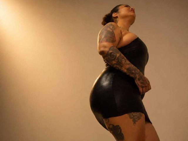 EsmeraldaStrong, com braços e coxas tatuados, vestindo um vestido preto de látex justo, está de costas para a câmera, olhando para cima, exibindo suas curvas e seu traseiro brilhante..