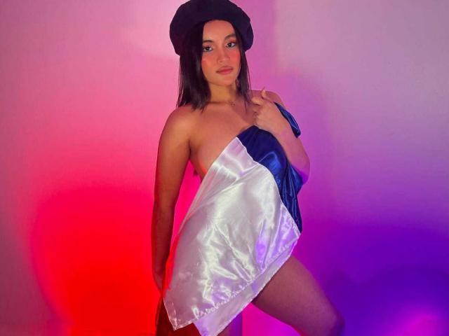 MiiaPalmer, avec une poitrine de taille moyenne, portant un béret noir et une robe blanche et bleue brillante, cachant ses seins avec une main, debout sous un éclairage violet et rouge..