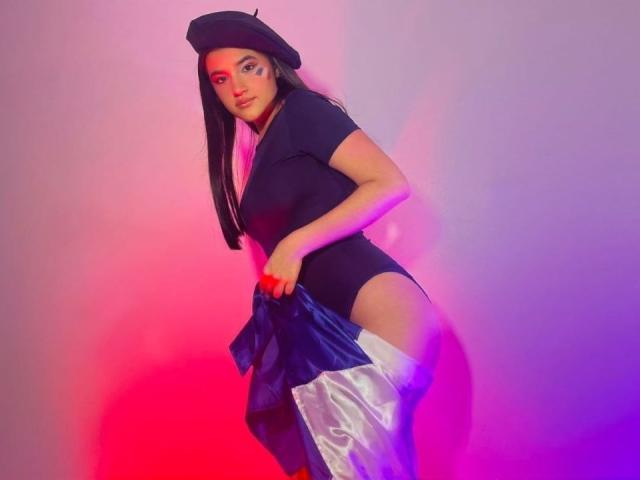 Iris Blair, met een zwarte hoed en een strak zwart top, houdt een blauw-witte rok vast, waardoor haar achterwerk zichtbaar is, voor een achtergrond die roze en paars is verlicht..
