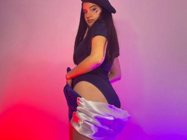 Iris Blair, met lang zwart haar, draagt een zwarte muts en een strak zwart topje, trekt zilveren satijnpantalon naar beneden om haar billen te laten zien, onder roze en paarse verlichting..