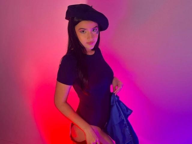 Iris Blair draagt een zwarte hoed en een strak shirt, ze houdt een blauw jack vast, met roze en rode verlichting, waarbij haar achterwerk zichtbaar is en ze naar de camera kijkt..