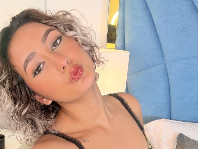 Elena Saenz, con i capelli ricci e dalle punte argentate, labbra rosa carnose e sopracciglia arcuate, indossa una camicetta di pizzo nero e guarda seducentemente l'obiettivo. Sullo sfondo, una testiera blu..